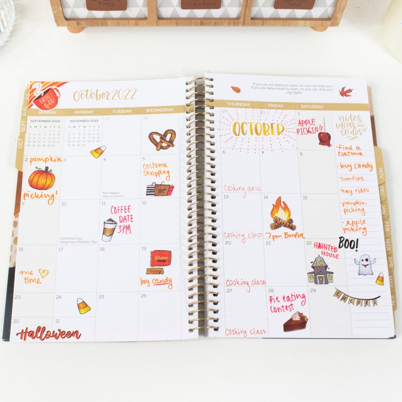 May include: A monthly planner with a gold spiral binding. The planner is open to October 2022. The pages are filled with handwritten notes and stickers with fall and Halloween themes. The stickers include images of pumpkins, candy corn, ghosts, and a haunted house.