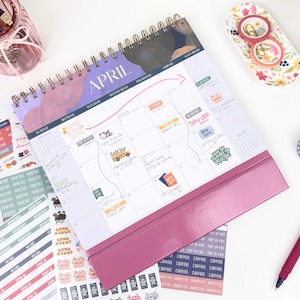 May include: A spiral-bound desk calendar for April with a purple and dark blue header. The grid features handwritten notes, colorful stickers, and a pink arrow. It includes a solid pink base and is surrounded by various sticker sheets and a pink pen.