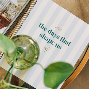 May include: A spiral-bound planner with a light blue and white vertical striped cover. The text reads 2027 the days that shape us with a small gold heart icon. A clear glass vase with green plant stems is positioned over the planner cover.