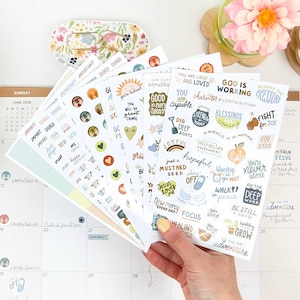 May include: A collection of adhesive sticker sheets featuring various inspirational phrases, small illustrations, and icons in a muted color palette. The designs include text such as God is working, You are capable, and Blessings are coming, printed on white paper.