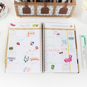 May include: An open planner with a June 2023 calendar page. The page is filled with colorful handwritten notes and drawings, including a watermelon, a beach ball, a camera, a flamingo, and a popsicle. The planner has a gold spiral binding.