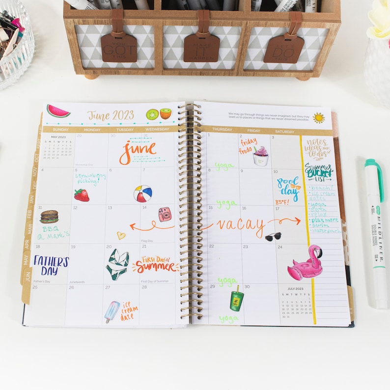 May include: An open planner with a June 2023 calendar page. The page is filled with colorful handwritten notes and drawings, including a watermelon, a beach ball, a camera, a flamingo, and a popsicle. The planner has a gold spiral binding.