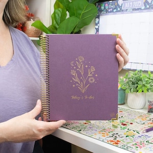 May include: A purple spiral-bound planner with gold accents and corner protectors. The cover features a gold floral design with the text "today's to-dos". The planner is held open, showcasing the design.
