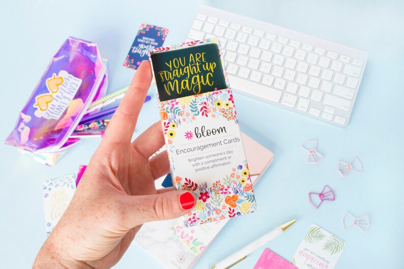 May include: A box of encouragement cards with a floral design. The box is open and the top card reads "You are straight up magic". The box is labeled "*bloom Encouragement Cards Brighten someone's day with a compliment or positive affirmation"