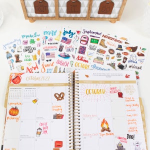 May include: A close-up of an open planner with a gold spiral binding. The planner is open to October 2022 and has stickers for each month of the year. The stickers are colorful and feature various designs, including pumpkins, a pretzel, a coffee cup, and a ghost.