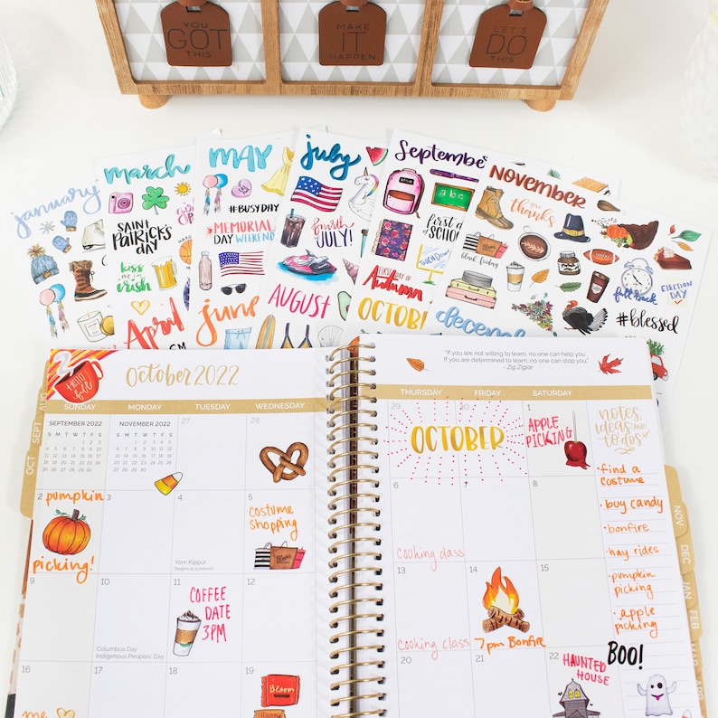 May include: A close-up of an open planner with a gold spiral binding. The planner is open to October 2022 and has stickers for each month of the year. The stickers are colorful and feature various designs, including pumpkins, a pretzel, a coffee cup, and a ghost.