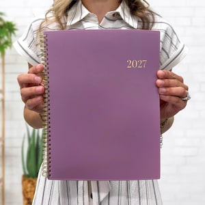 May include: A mauve-colored spiral-bound planner with the year 2027 printed in gold lettering on the front cover. The planner features a gold-toned metal wire binding along the left side and a smooth, solid-colored matte finish on the front cover.