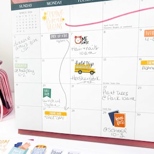 May include: A wall calendar for March 2026 featuring various colorful stickers and handwritten notes. The calendar includes labels such as Spring Break Begins, Pick Up Kids, Me Time, Field Trip, Half Day, Theme Day: Spirit Day, and Book Fair.
