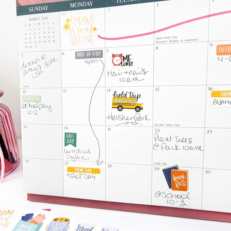 May include: A wall calendar for March 2026 featuring various colorful stickers and handwritten notes. The calendar includes labels such as Spring Break Begins, Pick Up Kids, Me Time, Field Trip, Half Day, Theme Day: Spirit Day, and Book Fair.