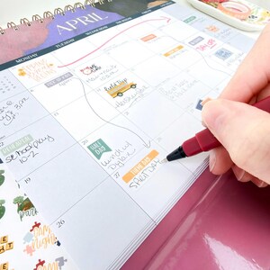 May include: A spiral-bound wall calendar for the month of April with handwritten notes and colorful stickers in the date squares. A dark red felt-tip pen is writing the words Spirit Day on the twenty-seventh square of the grid.
