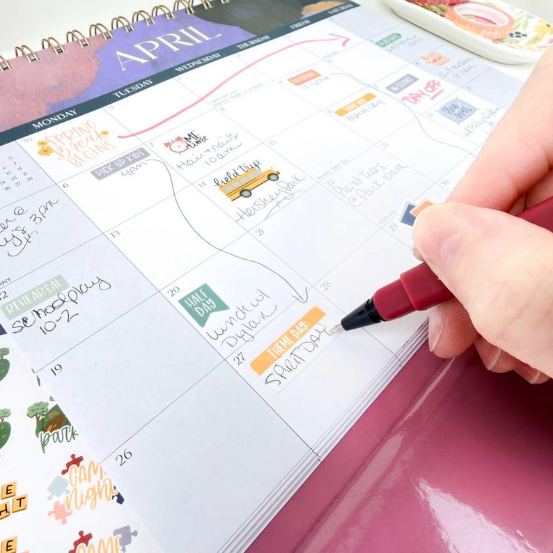May include: A spiral-bound wall calendar for the month of April with handwritten notes and colorful stickers in the date squares. A dark red felt-tip pen is writing the words Spirit Day on the twenty-seventh square of the grid.
