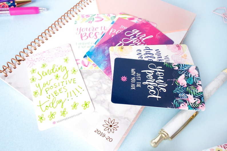 May include: A set of colorful affirmation cards with positive messages like "You're perfect just the way you are" and "Sending you positive vibes today!" The cards feature floral and flamingo designs.