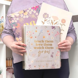 Op de afbeelding: Een verzameling bloemrijke lerarenplanners en notitieboekjes. De hoofdplanner is lichtgrijs met een gouden spiraalbinding en de tekst "teach them love them watch them grow". Andere items zijn een paars notitieboek met het nummer "26" en een crèmekleurige map.