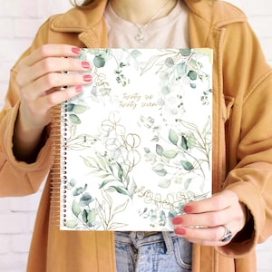May include: A spiral-bound planner with a white cover featuring a botanical design of green and gold leaves. The text "Twenty six Twenty seven" is printed in gold. The planner is held open, showcasing the design.