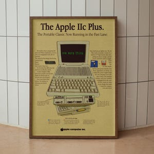 以下が含まれることがあります： Apple IIc Plusのコンピューターの額入りヴィンテージ広告。ポスターには、画面に「one more thing」というテキストが表示されたコンピューターのイラストが描かれています。ポスターは木製のフレームに入っています。