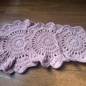 May include: Three handmade, light pink crochet coasters with a scalloped edge. The coasters are circular with a detailed, openwork design in the center. They are arranged on a wooden surface.