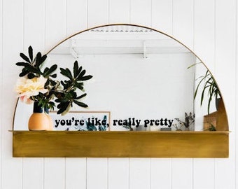 May include: A gold-colored shelf with a half-round mirror above it. The mirror reflects a plant in a vase and the text "you're like, really pretty" on a white background.