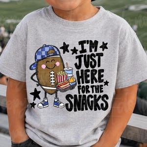 May include: A light gray short-sleeved t-shirt featuring a cartoon football character wearing a blue plaid cap and sneakers. The graphic includes a tray of snacks and the black text I'M JUST HERE FOR THE SNACKS surrounded by small star icons.