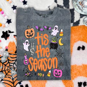 May include: A charcoal gray t-shirt featuring the orange text 'tis the season' surrounded by Halloween-themed graphics including a jack-o'-lantern, ghosts, a skeleton hand, a witch hat, a cauldron, a spider web, bats, and candy corn.