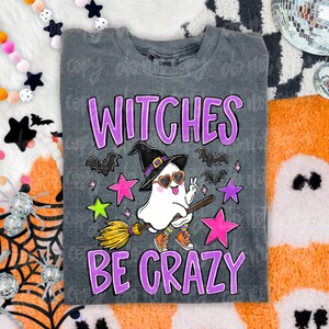 May include: A charcoal gray t-shirt featuring a graphic of a white ghost wearing a black witch hat and heart-shaped sunglasses, riding a broomstick. The text reads WITCHES BE CRAZY in purple letters, surrounded by pink, purple, and green stars.