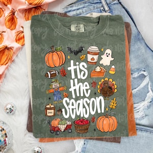 May include: An olive green t-shirt featuring the white text 'tis the season' surrounded by colorful illustrations of pumpkins, a scarecrow, a football, a bat, a coffee cup, a ghost, a slice of pie, a turkey, a candle, and a wagon of apples.