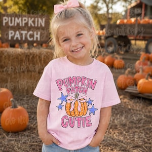 May include: A light pink short-sleeved t-shirt featuring a graphic of an orange pumpkin with a pink bow, surrounded by blue stars and the text PUMPKIN PATCH CUTIE in pink and black lettering. The shirt has a crew neckline and a casual fit.