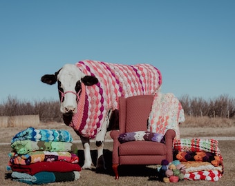 May include: A black and white cow wearing a pink, white, and red crocheted blanket. Beside the cow is a maroon armchair draped with a crocheted throw, surrounded by stacks of colorful, textured crocheted blankets and yarn balls.