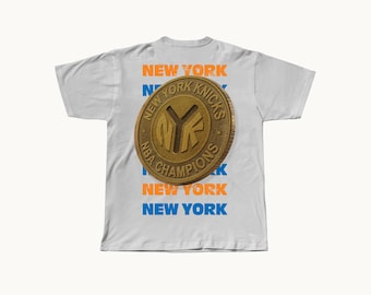 May include: A white short-sleeve t-shirt featuring a large gold-colored circular coin graphic in the center. The coin is embossed with the text NEW YORK KNICKS and NBA CHAMPIONS. Behind the coin, the words NEW YORK are printed in orange and blue.