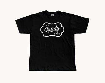 May include: A black short-sleeved t-shirt featuring a white graphic design on the chest. The design consists of a scalloped-edge frame containing the text 'I'M A' at the top, 'Grady' in a script font in the center, and 'BABY' at the bottom.