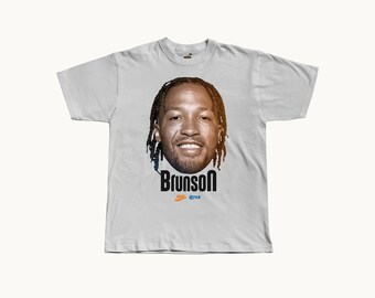 May include: A light gray short-sleeve t-shirt featuring a large photographic portrait of a smiling person with braided hair. Below the portrait, the text Brunson is printed in bold black letters, followed by a small orange Nike logo and Q718.