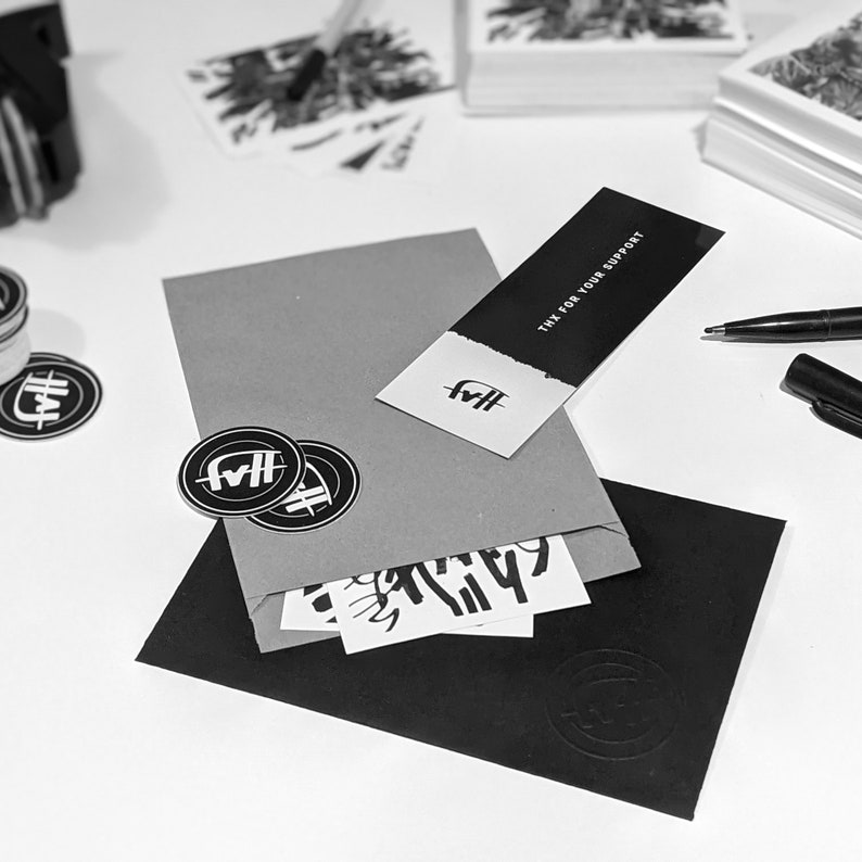 May include: A black and white sticker with the text "FWH" on a brown envelope. The envelope is on top of a black envelope. There is a white card with the text "Thx for your support" on the brown envelope. There are also other stickers on the table.
