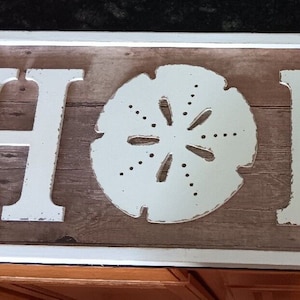 May include: White wooden sign with the word "HOME" spelled out in large, white letters. The letters are decorated with a seashell, a starfish, and a seashell.