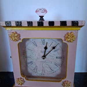 May include: A rectangular pink clock with a black and white striped top and bottom border. The clock face features Roman numerals, a cloud pattern, and the text Grand Central Terminal. It is adorned with four gold floral accents and a pink crystal knob.