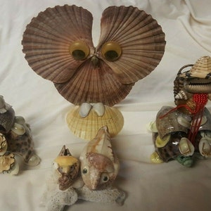 May include: Four handmade sculptures of animals made from seashells. The sculptures include an owl, a turtle, a crab, and a seahorse. The owl has large, round eyes and is made from a variety of seashells. The turtle is sitting on a base of seashells and has a small, round head. The crab is made from a variety of seashells and has a pair of large claws. The seahorse is made from a variety of seashells and has a long, thin neck.