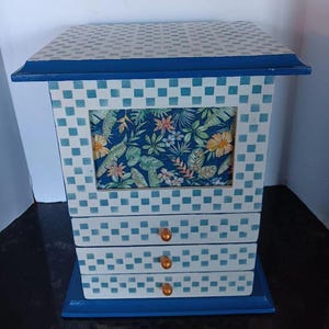 Puede incluir: Una caja de joyería de madera azul con un patrón de cuadros blancos y azules. La caja tiene tres cajones con pomos dorados y un panel con estampado floral en la parte delantera.