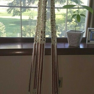 Puede incluir: Un macetero de macramé blanco y rojo con una maceta de vidrio verde colgando de él. El macetero está colgado frente a una ventana con vista a árboles y césped.