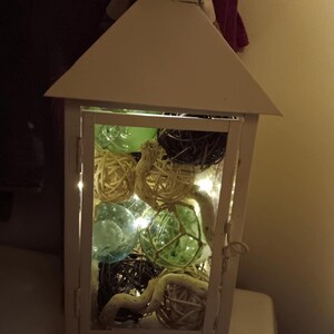Puede incluir: Linterna blanca con un panel de vidrio lleno de bolas decorativas blancas, marrones y verdes y una cadena de luces blancas.