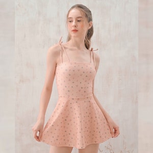 Pode incluir: Um vestido cor de pêssego com decote quadrado e alças finas amarradas nos ombros. O vestido apresenta um corpete justo e uma saia rodada, adornada com um delicado padrão floral em cinza suave. É um vestido de estilo de verão ou primavera.