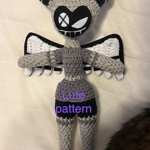 Puede incluir: Una muñeca de crochet gris y negro con alas, cuernos y una cara en blanco y negro. La muñeca lleva una falda a rayas grises y negras y tiene el texto "Lute pattern" escrito en ella.