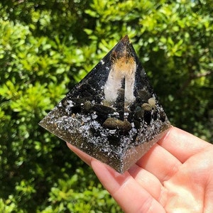 May include: A black pyramid-shaped crystal with embedded stones and glitter. The crystal is clear and has a light brown color in the center.