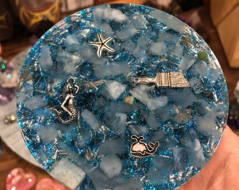 May include: Round, blue resin coaster with embedded blue stones and glitter. Silver charms of a starfish, mermaid, paint brush, and whale are set within the resin. A unique, decorative item.