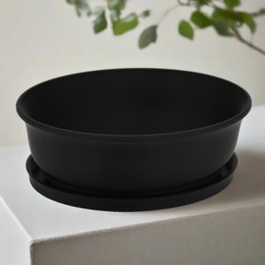 May include: A black plastic planter with a matching saucer. The planter is oval shaped and has a smooth, glossy finish.