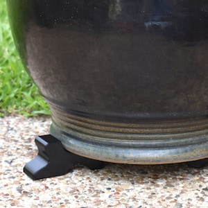 May include: A large, dark-colored ceramic planter with a glossy finish. The planter sits on a black, decorative stand with a curved design. The planter is on a stone surface with green grass in the background.