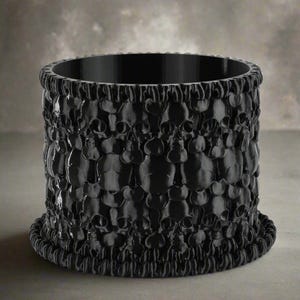 May include: A black cylindrical container with a textured surface. The container is covered in a repeating pattern of skulls. The top and bottom rims have a decorative, rope-like design. The container is made of a solid, dark material.