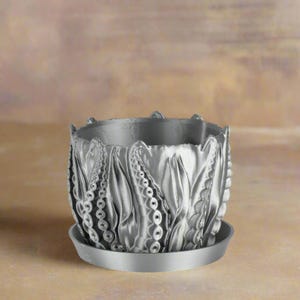 May include: A silver planter with a decorative, textured design resembling leaves or petals. The planter has a matching saucer.