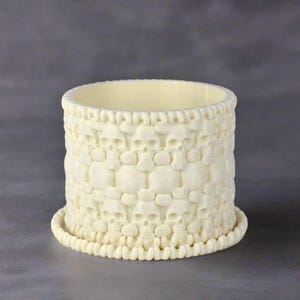 May include: White ceramic planter with a skull design. The planter has a wide, round base and a cylindrical body. The skull design is repeated around the entire planter.