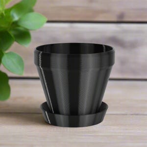 May include: A black plastic plant pot with a matching saucer. The pot has a smooth, slightly textured surface.