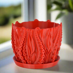 May include: A red ceramic planter with a unique design featuring an octopus-like pattern. The planter has a matching saucer.