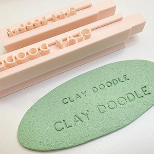 Könnte beinhalten: Zwei hellrosa rechteckige Tonformen mit dem Text "CLAY DOODLE" und ein grünes ovales Tonstück mit demselben Text. Die Formen werden verwendet, um Abdrücke in Ton zu erstellen.
