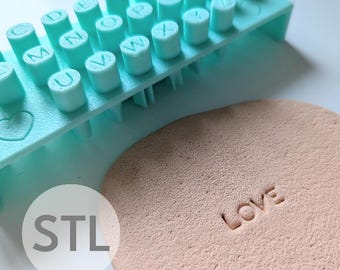 May include: A light blue plastic alphabet stamp set with 6mm letters. The set includes a heart stamp and a base with a handle. The stamp set is used to create the word "LOVE" on a beige surface.